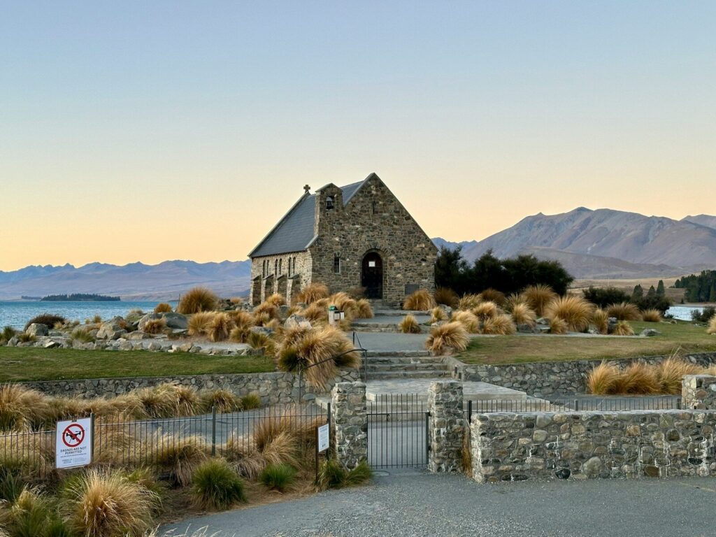 Lake Tekapo, New Zealand - Best Travel and Cruise New Zealand a stone church with a steeple surrounded by grass and mountains