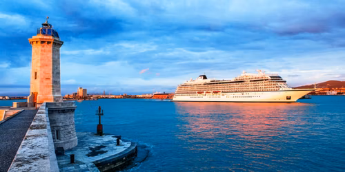 Lighthouse at a pier with a large cruise ship anchored in a calm harbor at sunset.