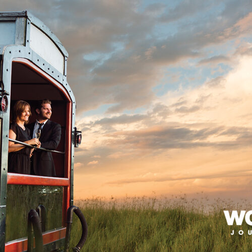 Couple in a vintage train carriage toasting a drink at sunset, with a grassy landscape in the background and a World Journeys logo in the corner.