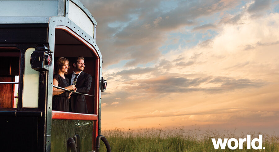 Couple in a vintage train carriage toasting a drink at sunset, with a grassy landscape in the background and a World Journeys logo in the corner.