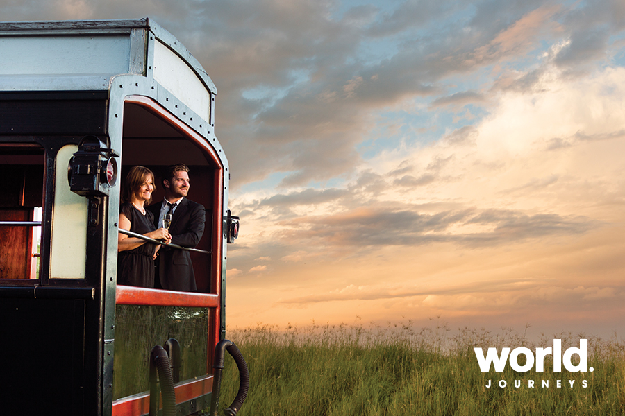Couple in a vintage train carriage toasting a drink at sunset, with a grassy landscape in the background and a World Journeys logo in the corner.
